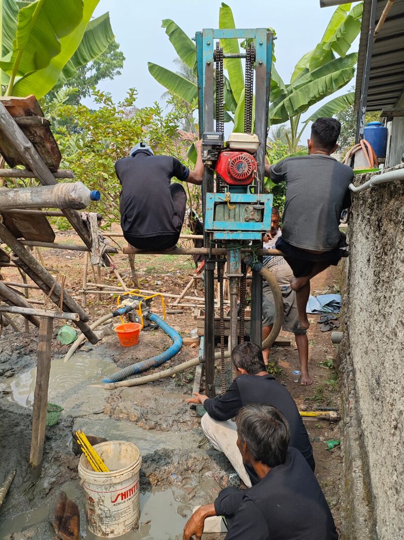 Tim Kenzie Sumur Bor mengoperasikan rig bor portabel di halaman rumah pelanggan, pipa sirkulasi air dan pompa terlihat di sekitar titik bor
